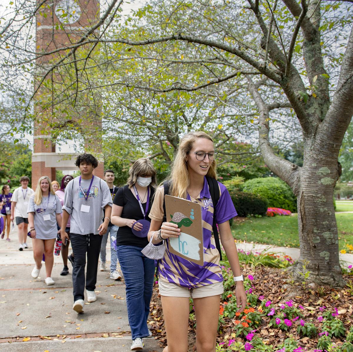 Group of college students walking on campus path under large trees holding clipboards and wearing lanyards.
