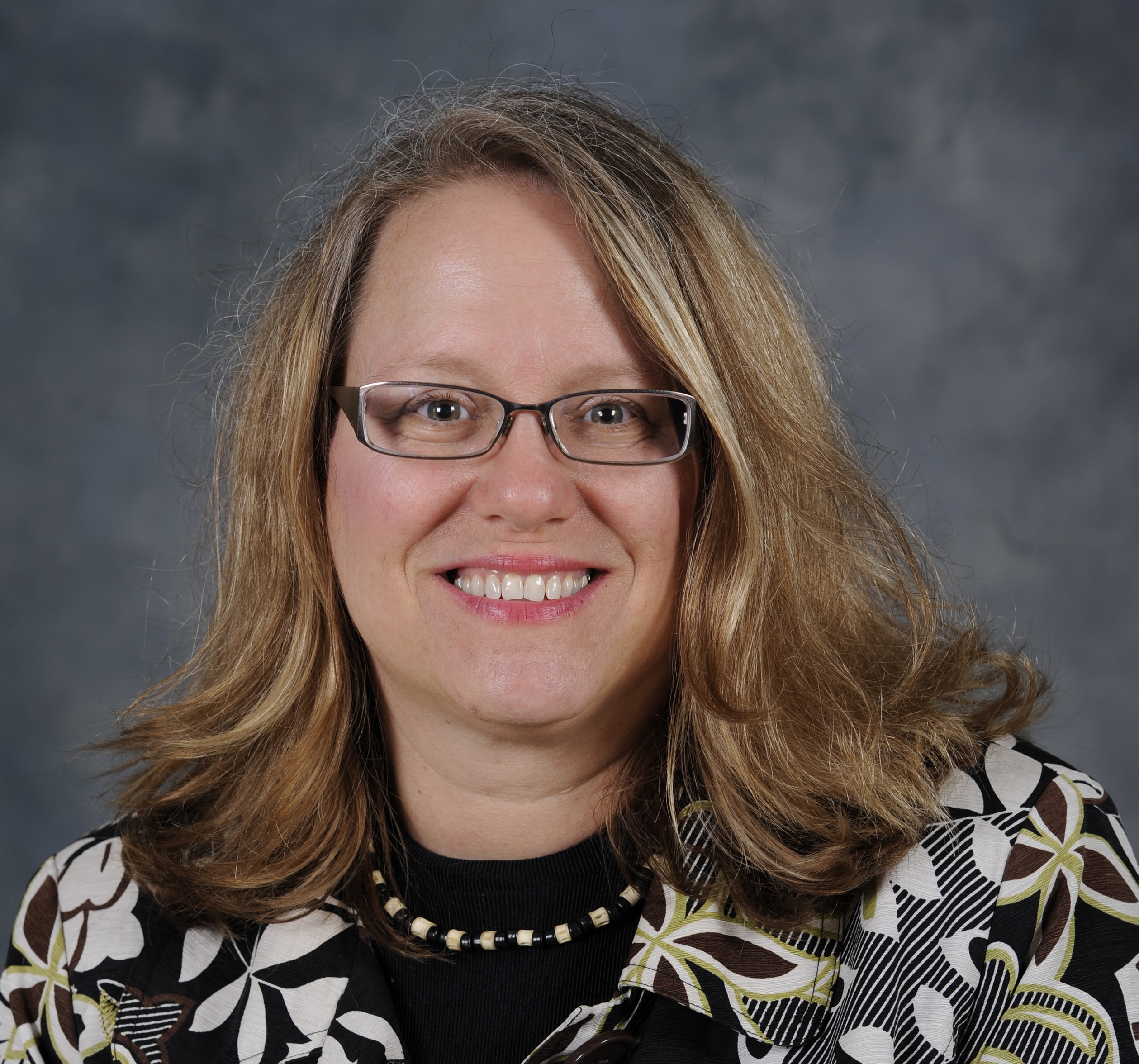 Photo of Barbara Jo White. A woman with light brown hair and glasses wearing a black and white blazer smiles with a dark blue backdrop behind her