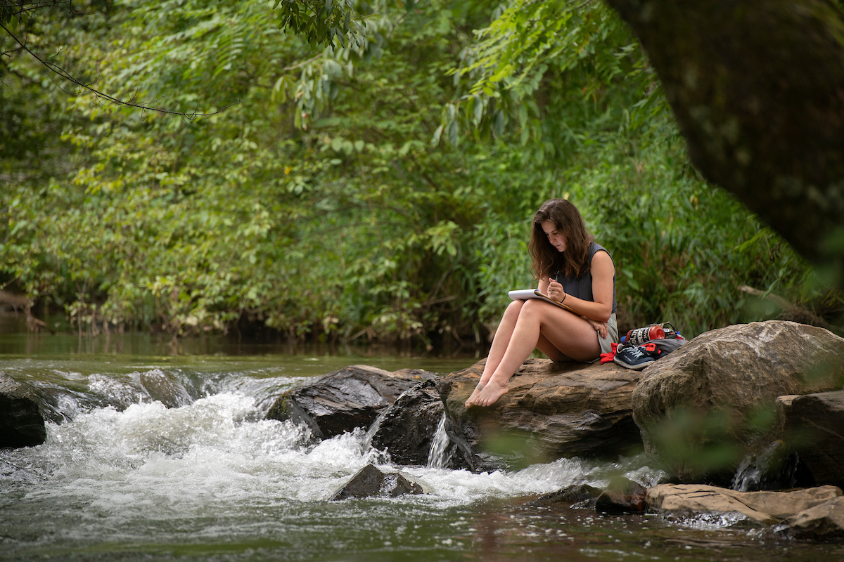 Student sits on a rock by a river and takes notes on a notepad