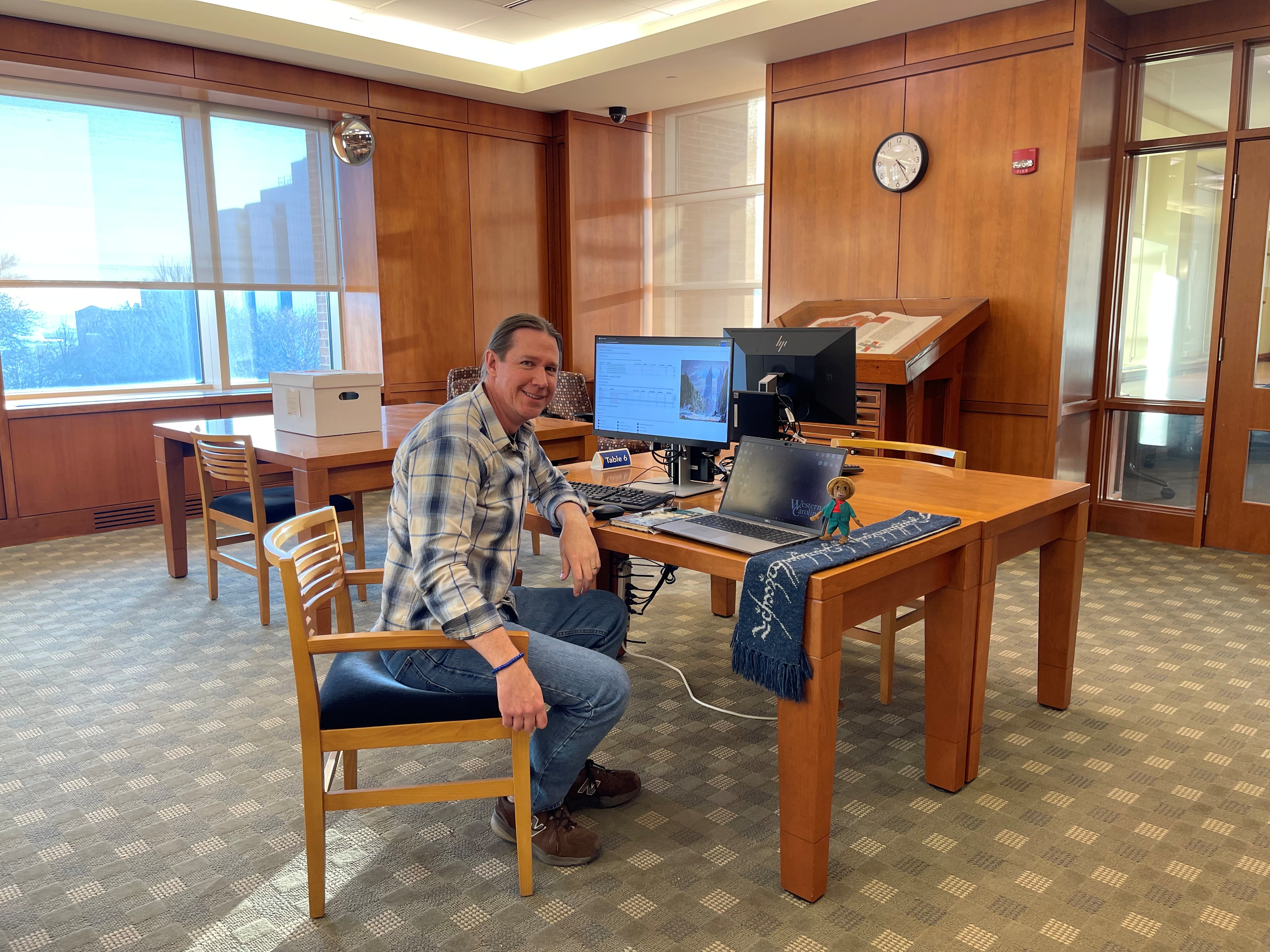 WCU professor John Whitmire sits in the Raynor Library at Marquette University, smiling while sitting in front of a laptop with a scarf and stuffed monkey beside the laptop